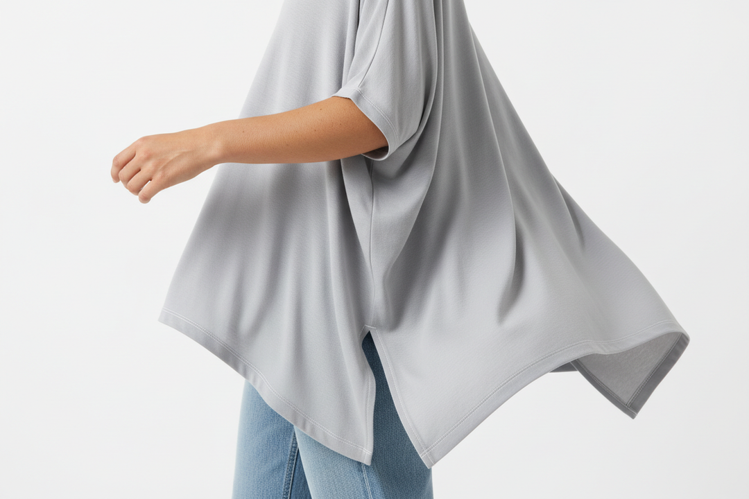 Close-up fashion detail shot of a woman wearing a loose, flowy light-gray top with an asymmetrical hem and side slit. Soft, lightweight fabric with natural folds and drape. Paired with light-wash blue jeans. Cropped from shoulder to hip, focus on garment movement and stitching details. Natural skin texture visible on the arm. Bright soft studio lighting, clean white background. Minimal, modern casual fashion aesthetic. High-resolution, photorealistic