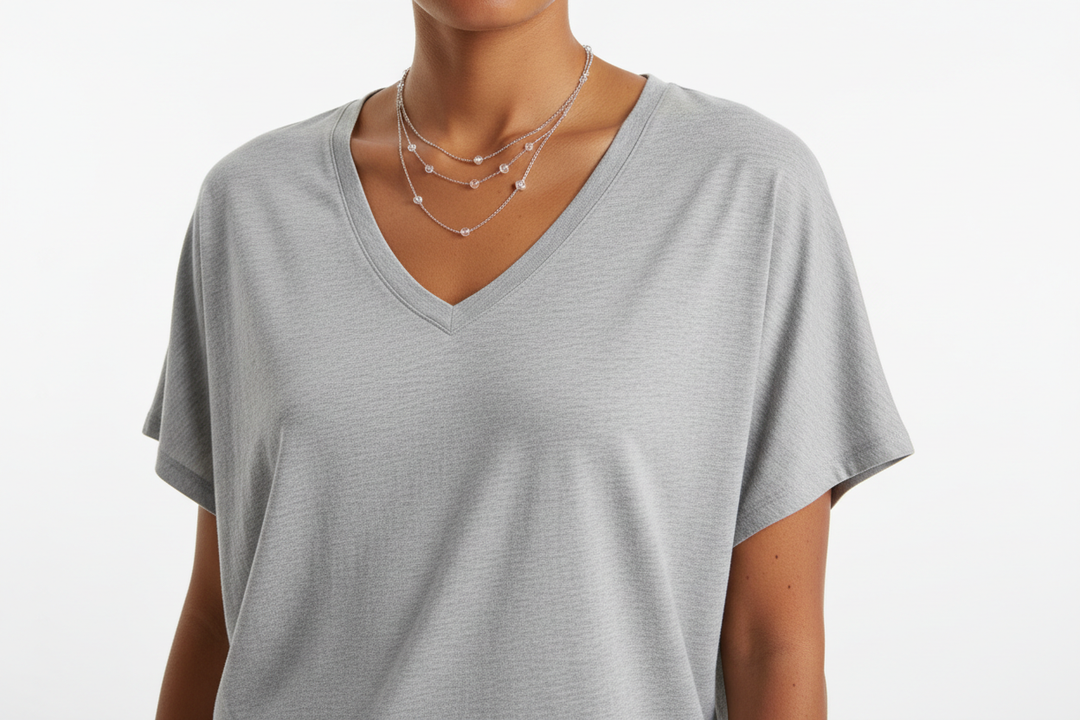 Close-up fashion photograph of a woman wearing a loose-fitting heather gray V-neck T-shirt with short sleeves. The fabric appears soft and lightweight with a relaxed drape. Minimalist style, neutral color palette. The frame shows the upper torso and shoulder area only, with natural skin tones and subtle freckles visible on the arm. Simple layered necklace with small beads. Bright, soft studio lighting on a clean white background. Modern casual apparel aesthetic, high-resolution, realistic texture